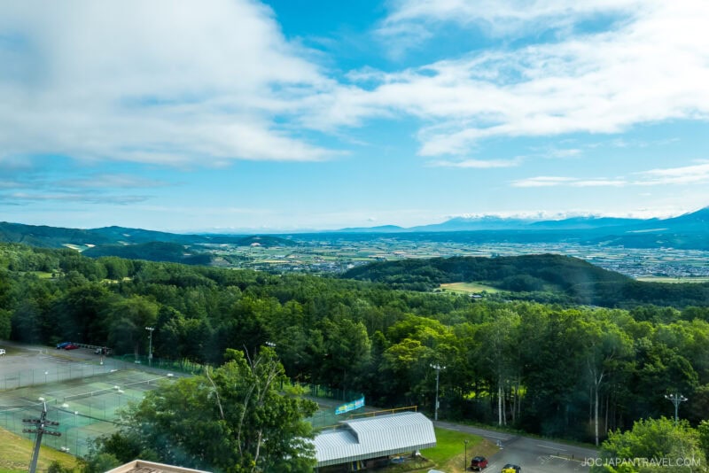 View of a lush green landscape with tennis courts and buildings in the foreground, dense forests, rolling hills, and an expansive blue sky with scattered clouds&mdash;likely taken from the New Furano Prince Hotel&rsquo;s elevated vantage point.