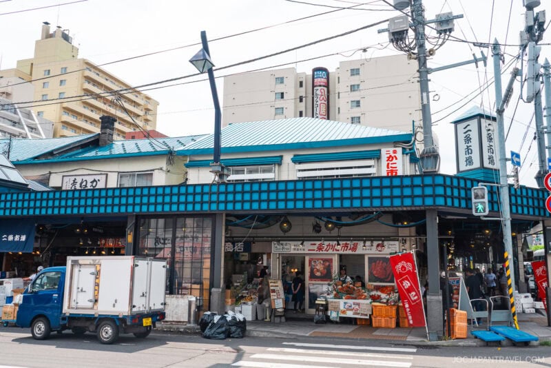 A busy street scene shows the blue-roofed Nijo Market building in Japan. Stalls display fruits, vegetables, and boxes, with people shopping and a small blue truck parked in front. Signs in Japanese hang above the entrance.