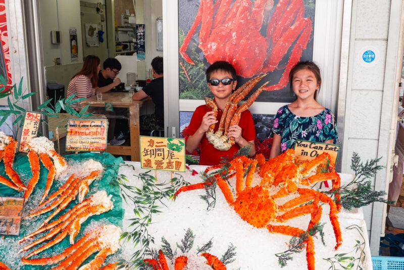 Two children stand in front of a seafood display at Nijo Market, one holding up a giant crab leg. Signs advertise crab dishes as people dine inside the bustling shop in the background.