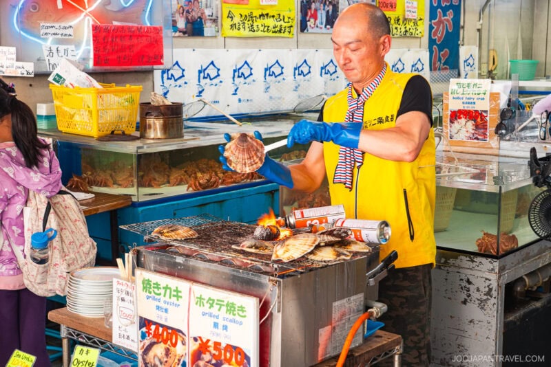 At Nijo Market, a man in a yellow vest grills scallops over an open flame at his seafood stall, surrounded by Japanese signs and fresh catch. Nearby, a customer with a backpack watches the cooking process.