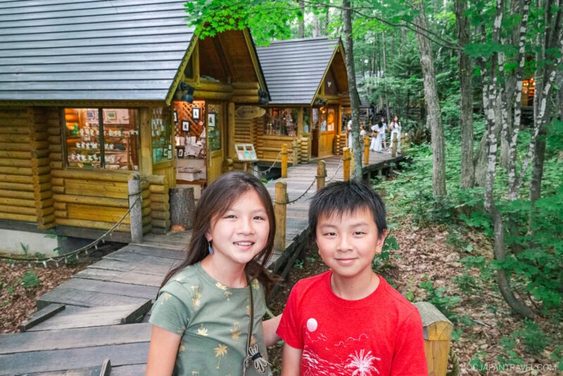 Two smiling children stand on the wooden path at Ningle Terrace, surrounded by lush trees and rustic log cabins with lit windows and display shelves. More people walk along the path in the background.