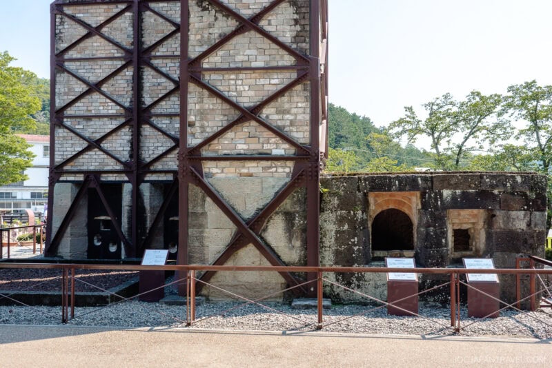 Two historic brick and stone furnace structures with metal supports are displayed outdoors at the Nirayama Reverberatory Furnace site, separated by a fence, with informational signs in front and trees and buildings in the background.