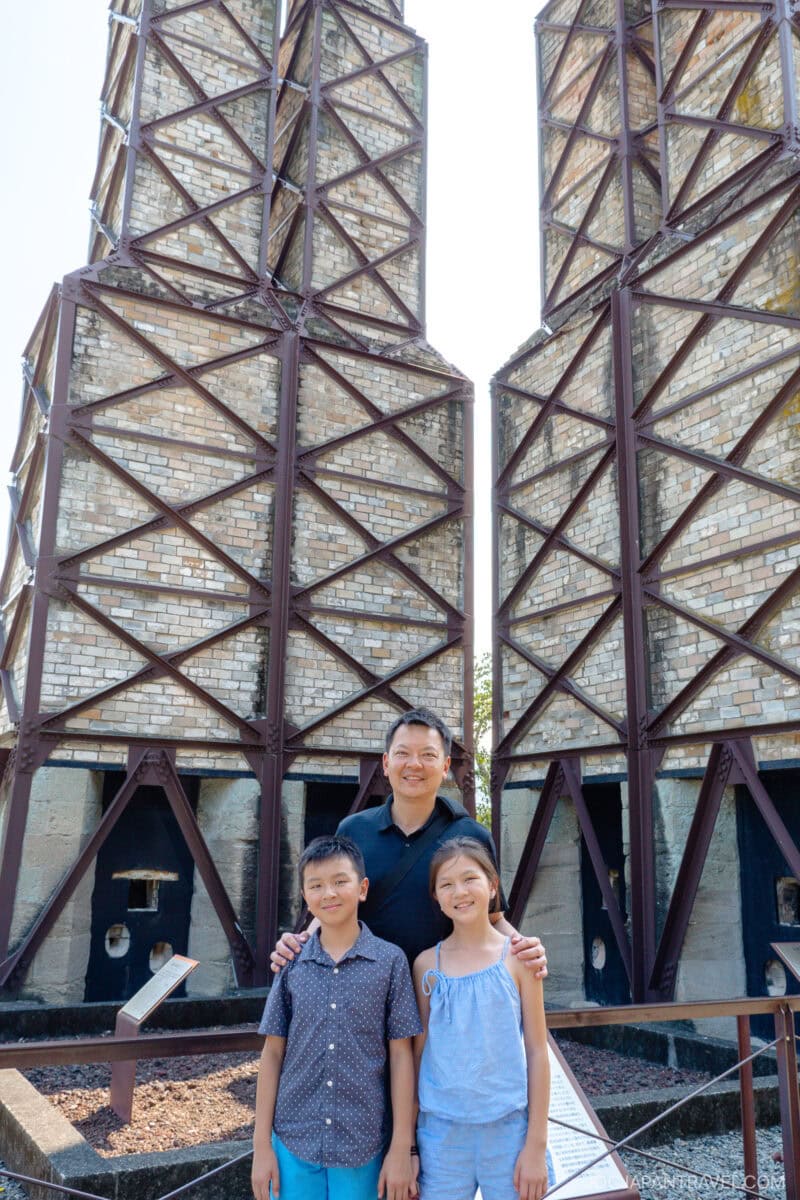 A man stands smiling with his hands on the shoulders of a boy and a girl in front of the historic Nirayama Reverberatory Furnace’s tall, brick industrial structures and metal frameworks, outdoors on a sunny day.