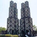 Nirayama Reverberatory Furnace(韮山反射炉) – Izu’s Industrial Revolution Legacy Two tall, rectangular brick towers with black metal frames stand side by side outdoors at Izunokuni Panorama Park, surrounded by greenery and a few visitors walking nearby on a sunny day.