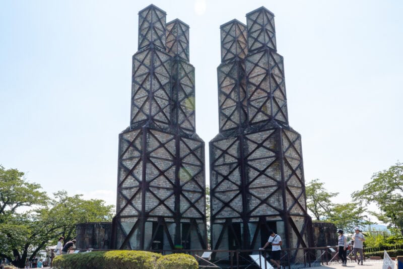 Two tall, narrow, brick towers with metal frameworks of the Nirayama Reverberatory Furnace stand side by side outdoors, surrounded by green trees and blue sky. Several people are walking and standing near the historic structures.