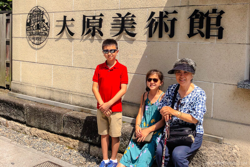 A boy in a red shirt, a girl in a blue dress, and an older woman in a patterned hat sit and stand outside the Ohara Museum of Art, with Japanese characters and a logo visible on the wall behind them.