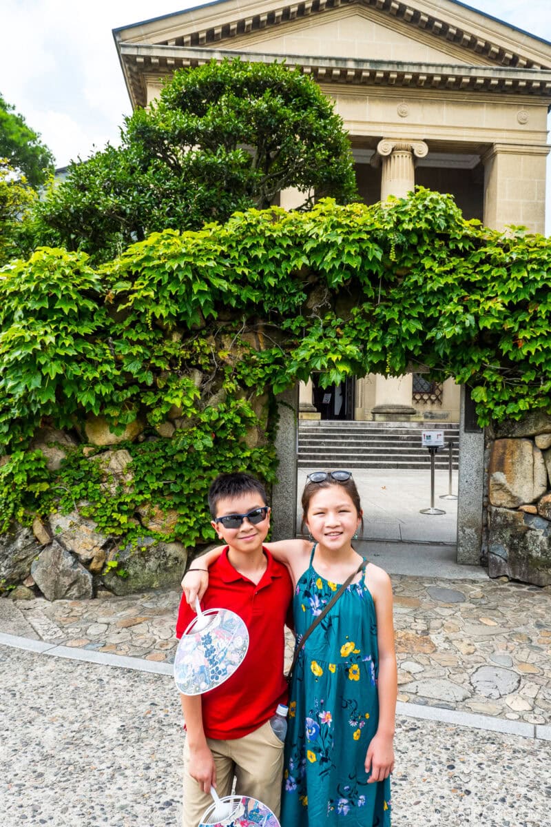 Two children, a boy in a red shirt and sunglasses and a girl in a green dress, stand smiling with arms around each other in front of a vine-covered stone archway near the Ohara Museum of Art’s classical columned building.