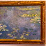 A framed painting of Claude Monet’s Water Lilies, displayed on a gallery wall at the Ohara Museum of Art, shows lily pads and flowers floating on a blue-green pond, with subtle reflections and soft, impressionist brushstrokes.