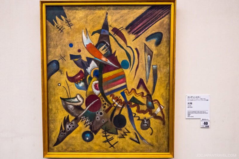 A colorful abstract painting with geometric shapes, bold lines, and vibrant patterns on a yellow background is framed in gold, displayed on a gallery wall at the Ohara Museum of Art with an information plaque beside it.