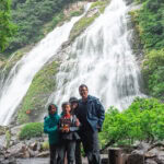 Three people in rain jackets stand together in front of Ohko Waterfall, its powerful cascades surrounded by lush green trees and rocky terrain. Mist from the waterfall fills the background.