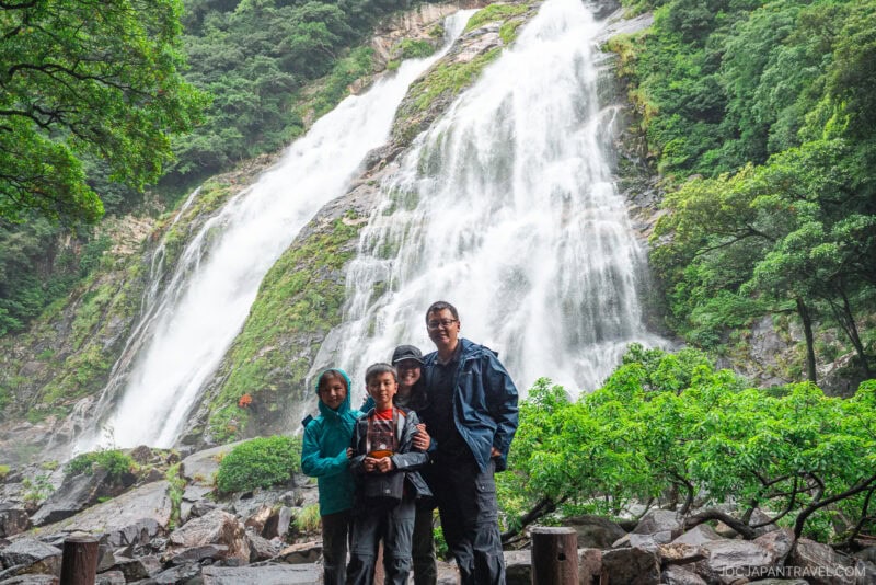 Three people in rain jackets stand together in front of Ohko Waterfall, its powerful cascades surrounded by lush green trees and rocky terrain. Mist from the waterfall fills the background.