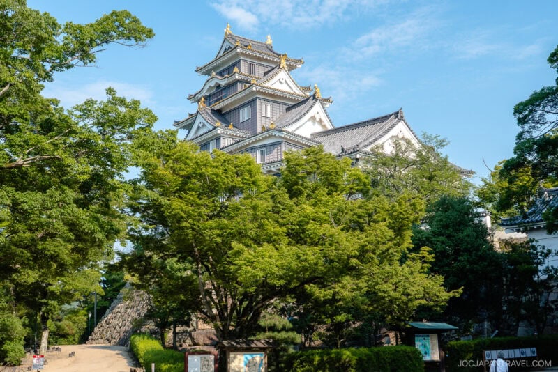 Okayama Castle, a traditional Japanese fortress with white walls and black tiled roofs, rises above lush green trees under a clear blue sky.