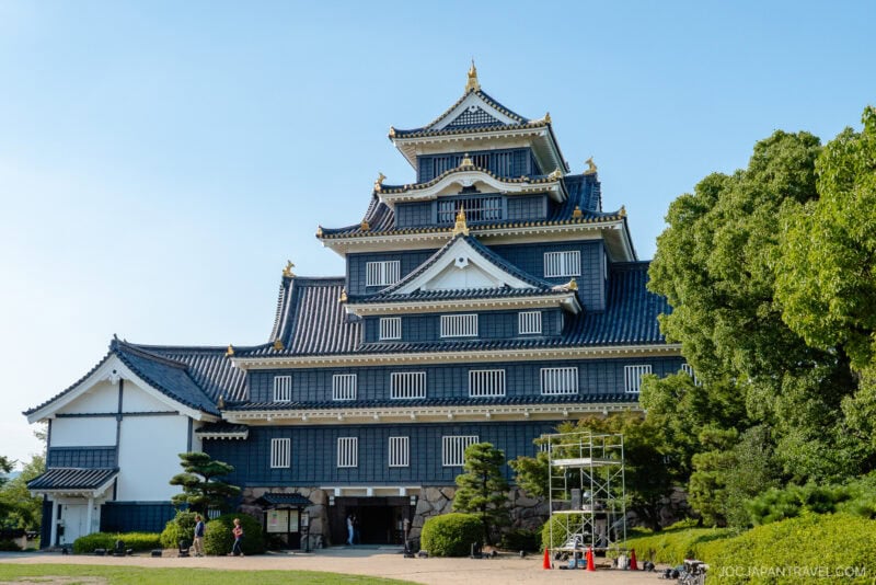 Okayama Castle, a traditional Japanese structure with black walls and gold accents, stands behind trees and scaffolding on a sunny day, set against a clear blue sky.