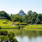 A serene Japanese garden, inspired by Okayama Korakuen Garden, features manicured trees, a pond, a small wooden bridge, and traditional huts with a white castle rising above the lush greenery under a clear blue sky.
