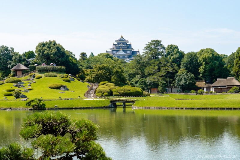 A serene Japanese garden, inspired by Okayama Korakuen Garden, features manicured trees, a pond, a small wooden bridge, and traditional huts with a white castle rising above the lush greenery under a clear blue sky.