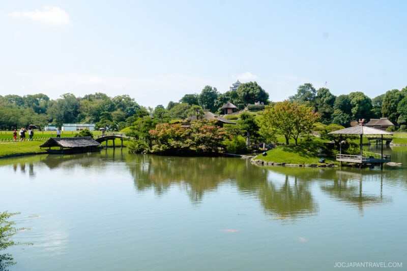 A serene Japanese garden reminiscent of Okayama Korakuen Garden, with a large pond, small wooden bridges, lush greenery, and a traditional gazebo, as people stroll in the distance beneath a clear blue sky.