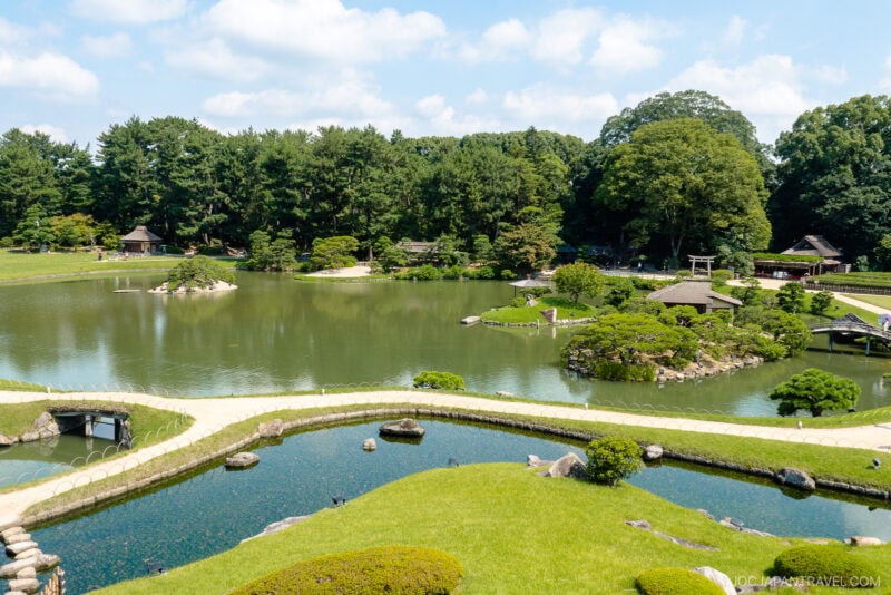 A traditional Japanese garden, Okayama Korakuen Garden features a large pond, curved stone bridges, manicured lawns, and lush green trees under a partly cloudy sky. Small wooden structures are seen around the water.
