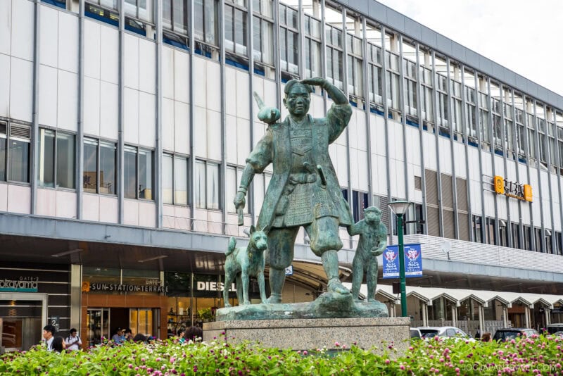 A bronze statue of a man in traditional clothing with animals by his side stands before a modern building with large windows in Okayama. Bushes with pink flowers bloom in the foreground near street-level storefronts.