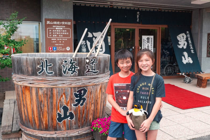 Two smiling children stand in front of a large wooden barrel with Japanese writing outside the Otokoyama Sake Museum; the boy wears a red shirt, and the girl in a dark shirt holds a stuffed penguin.