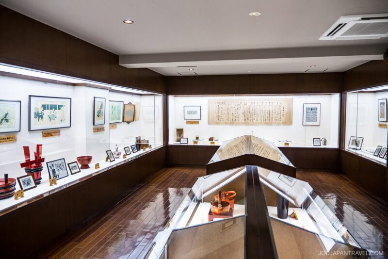 A museum exhibit room at the Otokoyama Sake Museum features glass display cases with artifacts, photographs, and framed artwork on the walls; the wooden floor and ceiling lights create a warm, inviting atmosphere.