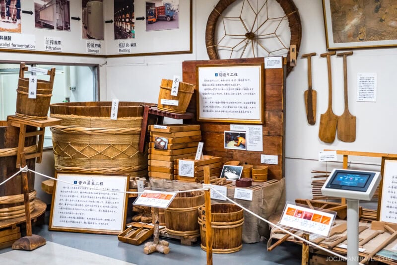The Otokoyama Sake Museum exhibit displays traditional Japanese wooden tools and containers for sake brewing, with labels and informational signs in Japanese, all showcased under bright lighting.