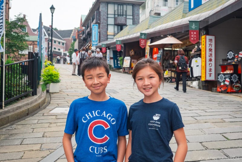 Two smiling children stand close together on a stone-paved street lined with shops and signs in Japanese, not far from Oura Church. The boy wears a blue Chicago Cubs shirt; the girl wears a blue shirt with white text. People walk in the background.