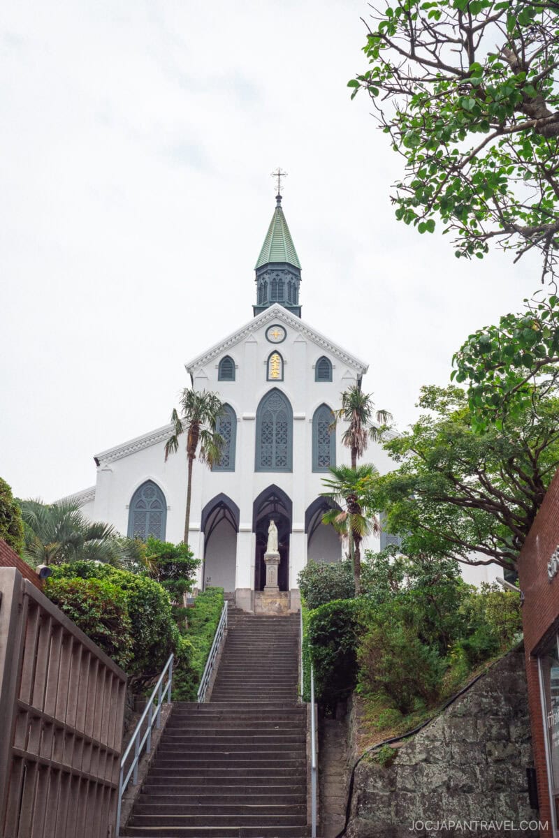 Oura Church, with its white facade, pointed arches, and green spire, stands at the top of a stone staircase surrounded by greenery. A statue near the entrance adds to the peaceful and inviting scene.