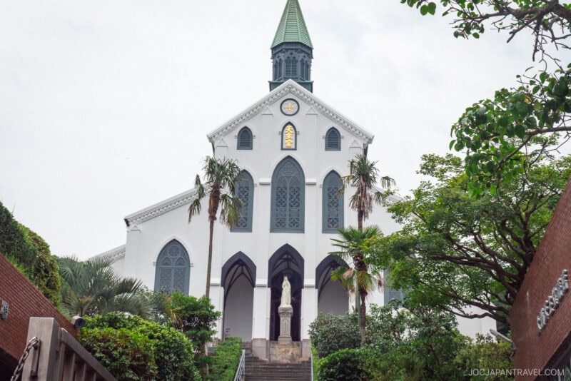 Oura Church, with its white fa&ccedil;ade, green steeple, and large arched windows, stands on a small hill surrounded by lush trees. A statue graces the center of stone steps leading to the entrance.