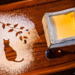 A wooden tray with a cat and branch design made from powdered sugar on the left, and a square ceramic cup of Tenshin-an custard dessert with a glossy spoon on the right.