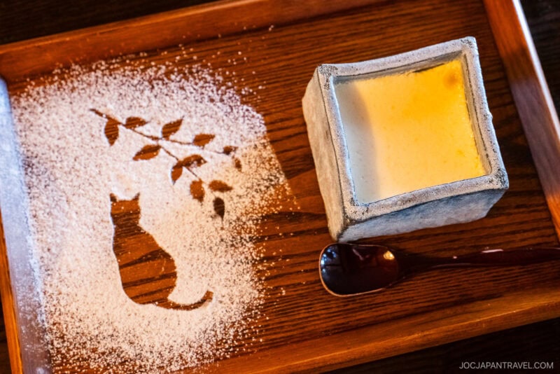 A wooden tray with a cat and branch design made from powdered sugar on the left, and a square ceramic cup of Tenshin-an custard dessert with a glossy spoon on the right.