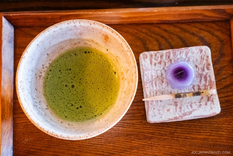 A bowl of frothy green matcha tea is served on a wooden tray at Tenshin-an, accompanied by a small purple wagashi sweet on a patterned plate with a wooden pick.