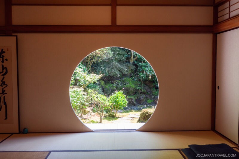 A circular window in the traditional Japanese tatami room of Tenshin-an frames a view of a lush garden with green foliage and rocks outside. Soft natural light fills the serene, minimalist interior.
