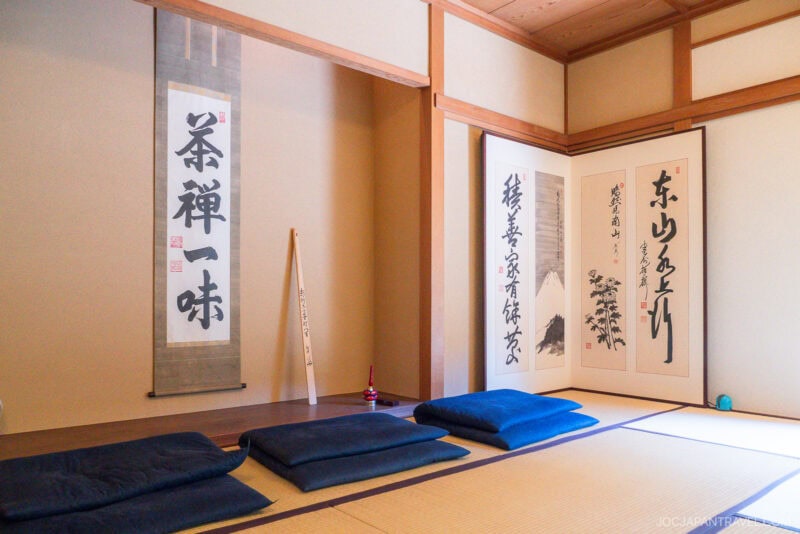 A traditional Japanese tatami room at Tenshin-an features blue floor cushions, scrolls with Japanese calligraphy on the wall, and a bamboo tea whisk in the corner, creating a serene atmosphere.