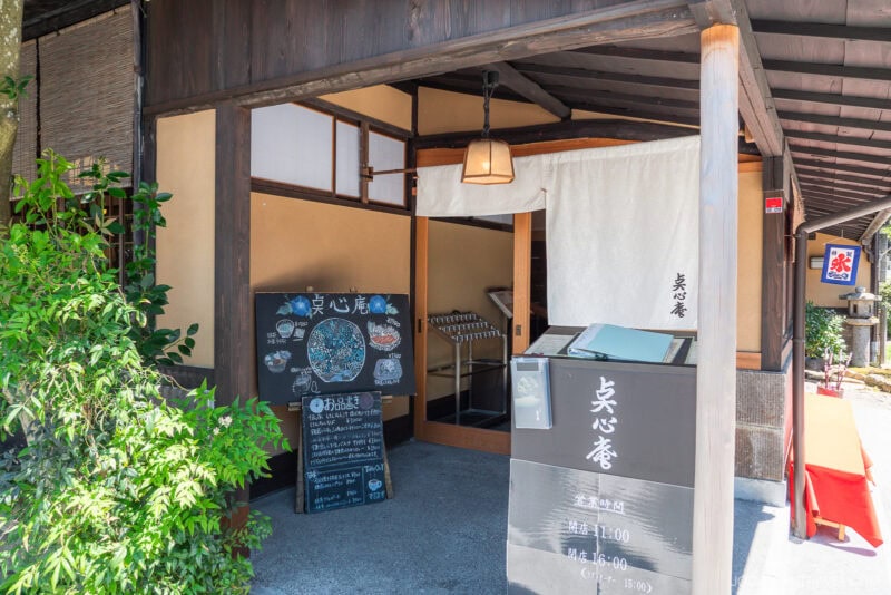 Tenshin-an’s traditional Japanese restaurant entrance features a wooden exterior, menu boards with dishes and information, a cloth curtain above the doorway, and greenery on the side. The setting appears calm and inviting.