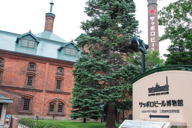 A historic red-brick building with a tall chimney labeled &ldquo;Sapporo Beer&rdquo; in Japanese; the Sapporo Beer Museum sign and lush green trees are in the foreground.
