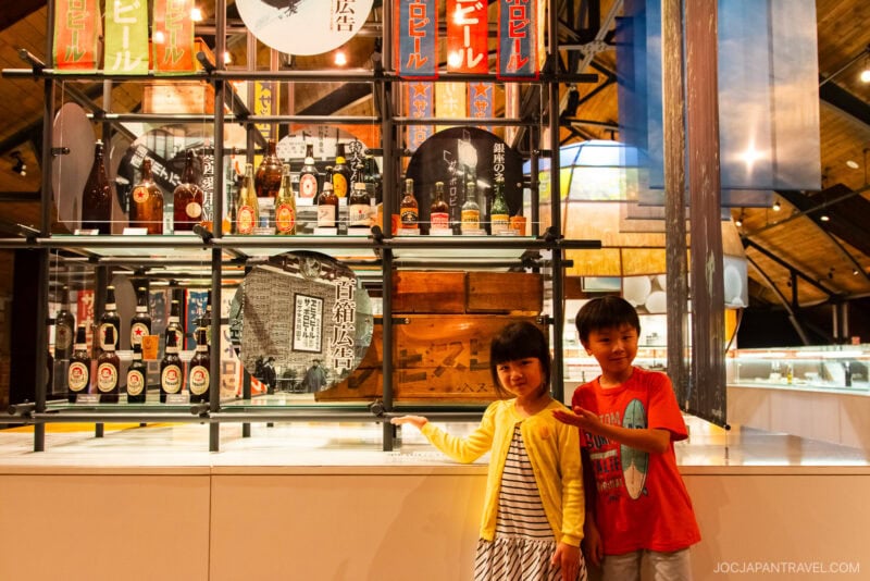 Two smiling children stand indoors, posing in front of a display of bottles and vintage Japanese signs at the Sapporo Beer Museum. The background features glass cases and warm lighting.