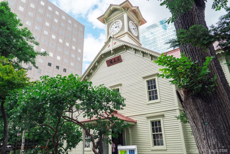 The Sapporo Clock Tower, a historic wooden building with a red roof, stands among trees and contrasts beautifully with surrounding modern glass and concrete buildings under a partly cloudy sky.