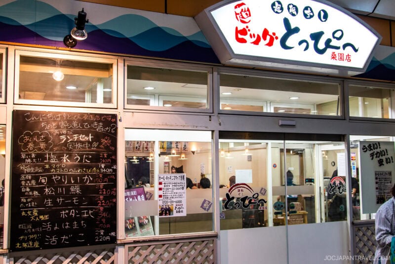 A Japanese restaurant exterior at night with large windows, an illuminated interior, a menu board with handwritten Japanese text, and a blue sign above the entrance. Inside, people are enjoying Conveyor Belt Sushi as they dine.