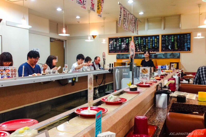 People sit at a conveyor belt sushi restaurant, selecting plates of fresh sushi as they pass by. The bright interior features Japanese menu signs and a staff member behind the counter, creating an authentic Conveyor Belt Sushi experience.
