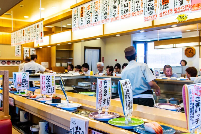 A busy Conveyor Belt Sushi restaurant with customers seated at the counter. Plates of sushi glide by, while chefs work behind the counter. Japanese menu signs hang above, displaying dish names and prices.