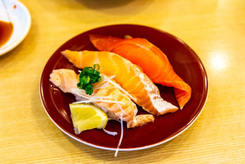 A red plate from a Conveyor Belt Sushi restaurant features assorted salmon sushi: two pieces of raw salmon nigiri and one seared salmon, topped with green onions, shredded radish, and a lemon wedge.