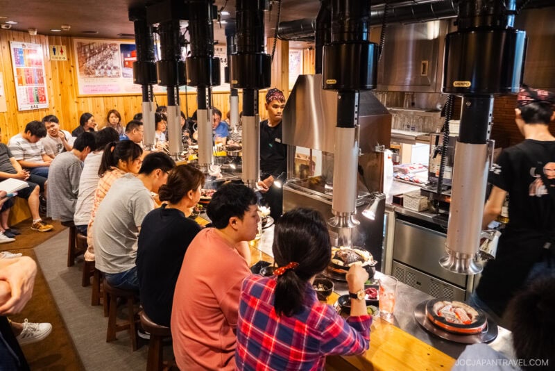 A busy Japanese restaurant with diners seated at long wooden tables, grilling Jingisukan on tabletop grills. The lively room buzzes with conversation and the aroma of grilled meat under large metal ventilation hoods.