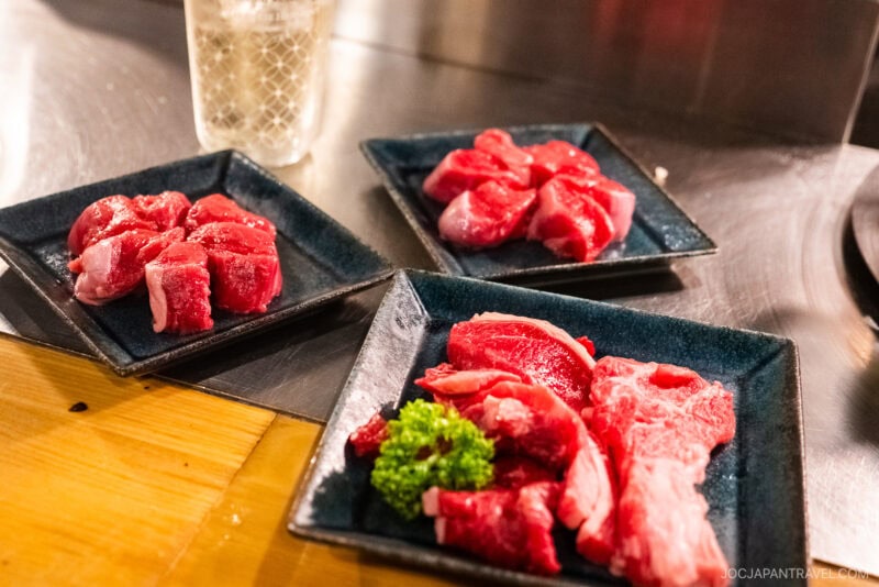 Three black plates with raw cuts of red meat for Jingisukan are displayed on a wooden counter, with a garnish of parsley on one plate and a glass of ice water in the background.