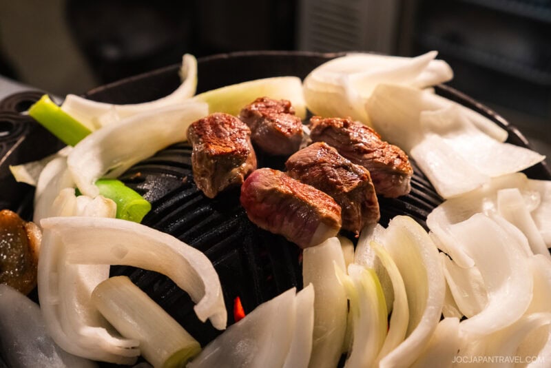 Thick slices of beef and rings of white onion grill on a round, black, ridged Jingisukan barbecue with visible flames underneath. The food is surrounded by more raw onion slices waiting to cook.