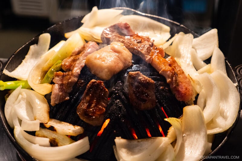 Slices of meat and onions are grilling on a round, black cast iron grill with red-hot charcoal underneath, creating smoke as the Jingisukan cooks.