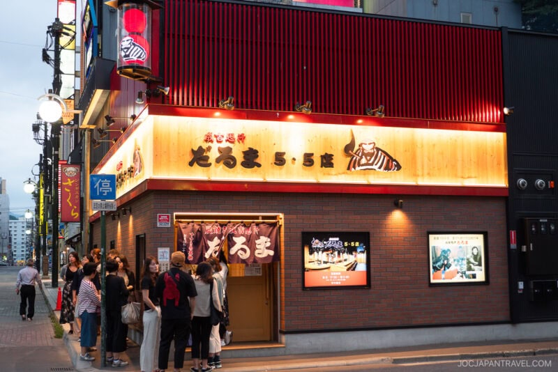 A group of people stand outside a brightly lit restaurant with Japanese signage and a cow illustration, known for its Jingisukan, located on a bustling city street in the evening.