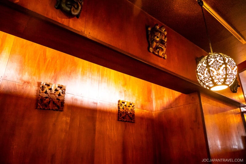 Warmly lit wooden wall with decorative carved masks and panels, and an ornate hanging lamp casting patterned shadows. The setting feels cozy and inviting—perfect for savoring Sapporo Soup Curry in a space with a traditional artistic touch.