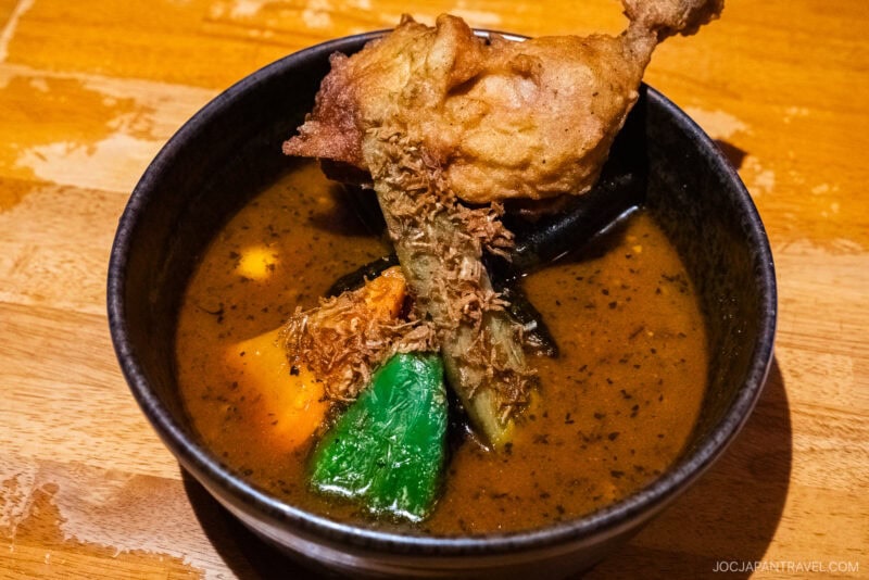 A bowl of Sapporo Soup Curry with a piece of fried chicken, leafy greens, and vegetables in a rich, brown broth, served on a wooden table.