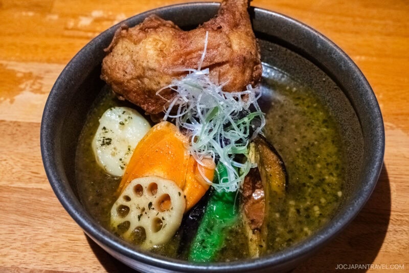 A bowl of Sapporo Soup Curry features a fried chicken leg, sliced carrots, lotus root, potatoes, eggplant, green pepper, and shredded white onions in a rich broth, served in a black bowl on a wooden table.
