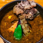 A bowl of Sapporo Soup Curry featuring tender pieces of meat, carrot, eggplant, green pepper, and other vegetables in a rich, dark broth, served in a black bowl on a wooden table.